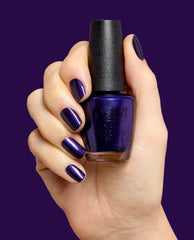 Hand with glossy deep purple nails holds an OPI Don't Over Ink It nail lacquer bottle against a purple background, showcasing a shimmering shade for fall nail colors.