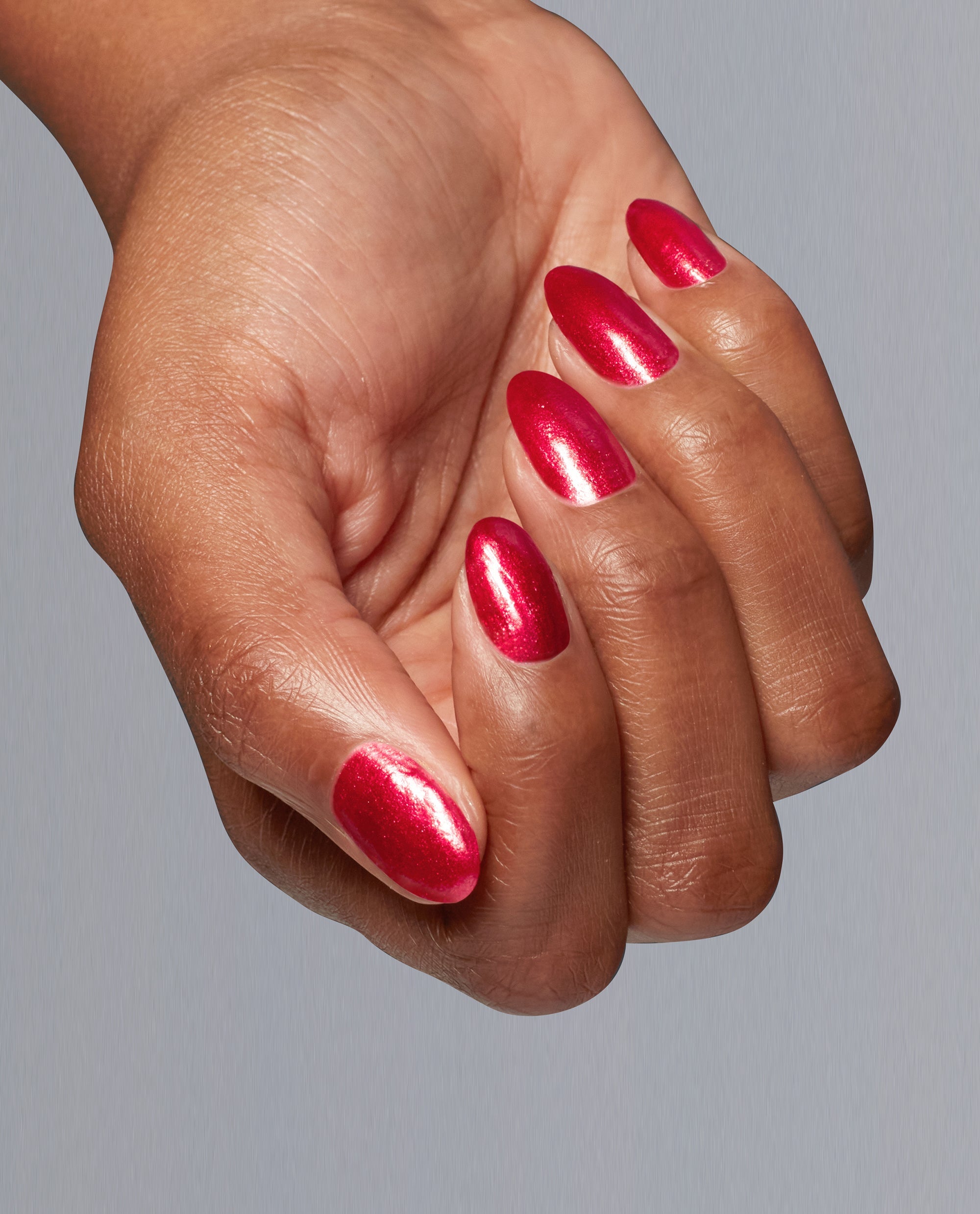 Hand with medium skin tone showing almond-shaped nails painted in Kiss My Aries, a bright shimmery red OPI nail polish lacquer, held against a smooth light gray background to highlight the bold glossy manicure color.