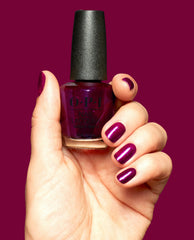 Hand with glossy deep berry-purple nails holds an OPI Lincoln Park After Brunch Nail Lacquer bottle, showing matching shimmer polish against a solid burgundy background.
