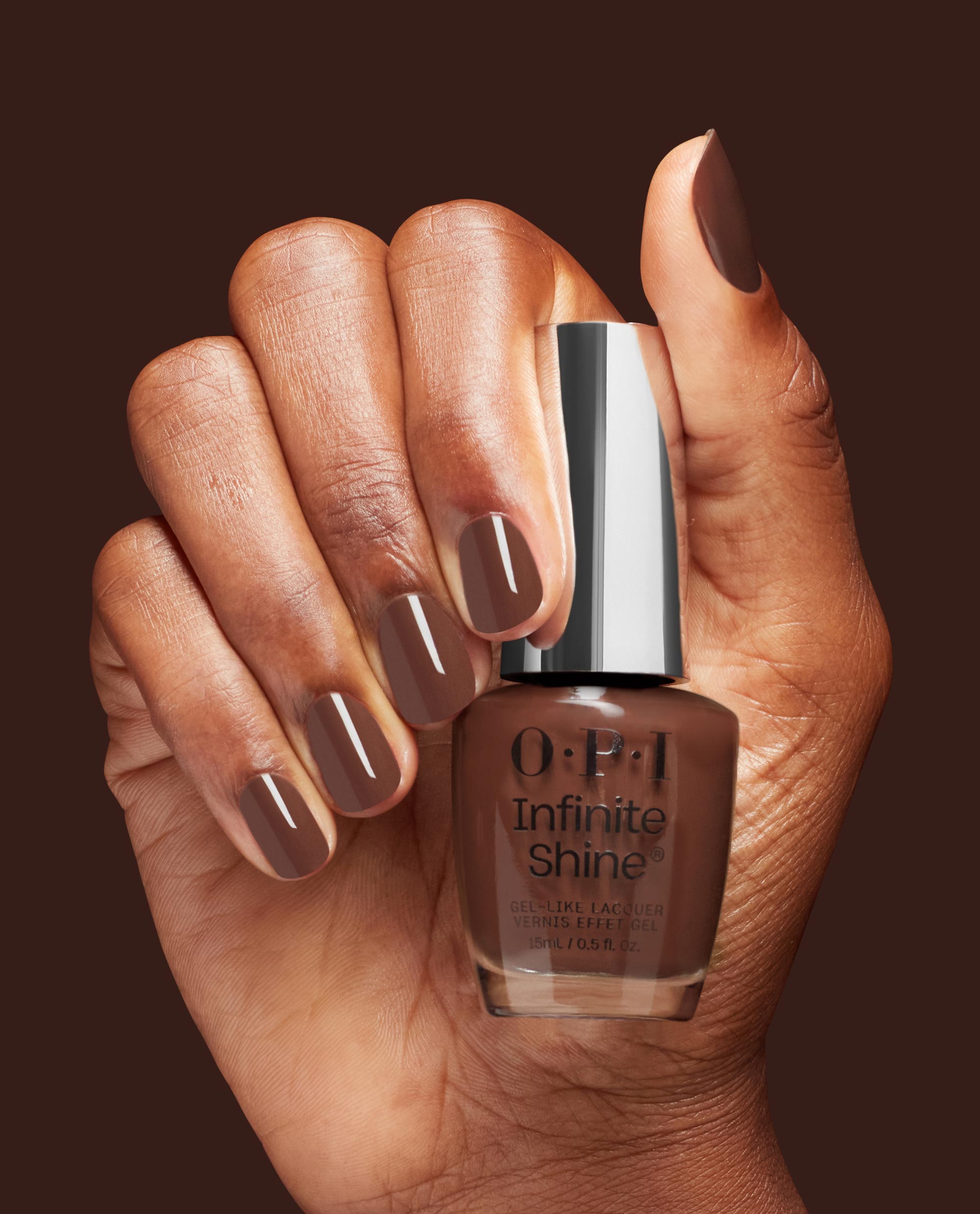 Hand with glossy brown manicured nails holds an OPI Infinite Shine Lip Hot Liner gel-like lacquer bottle against a dark brown background, showcasing warm fall nail colors.