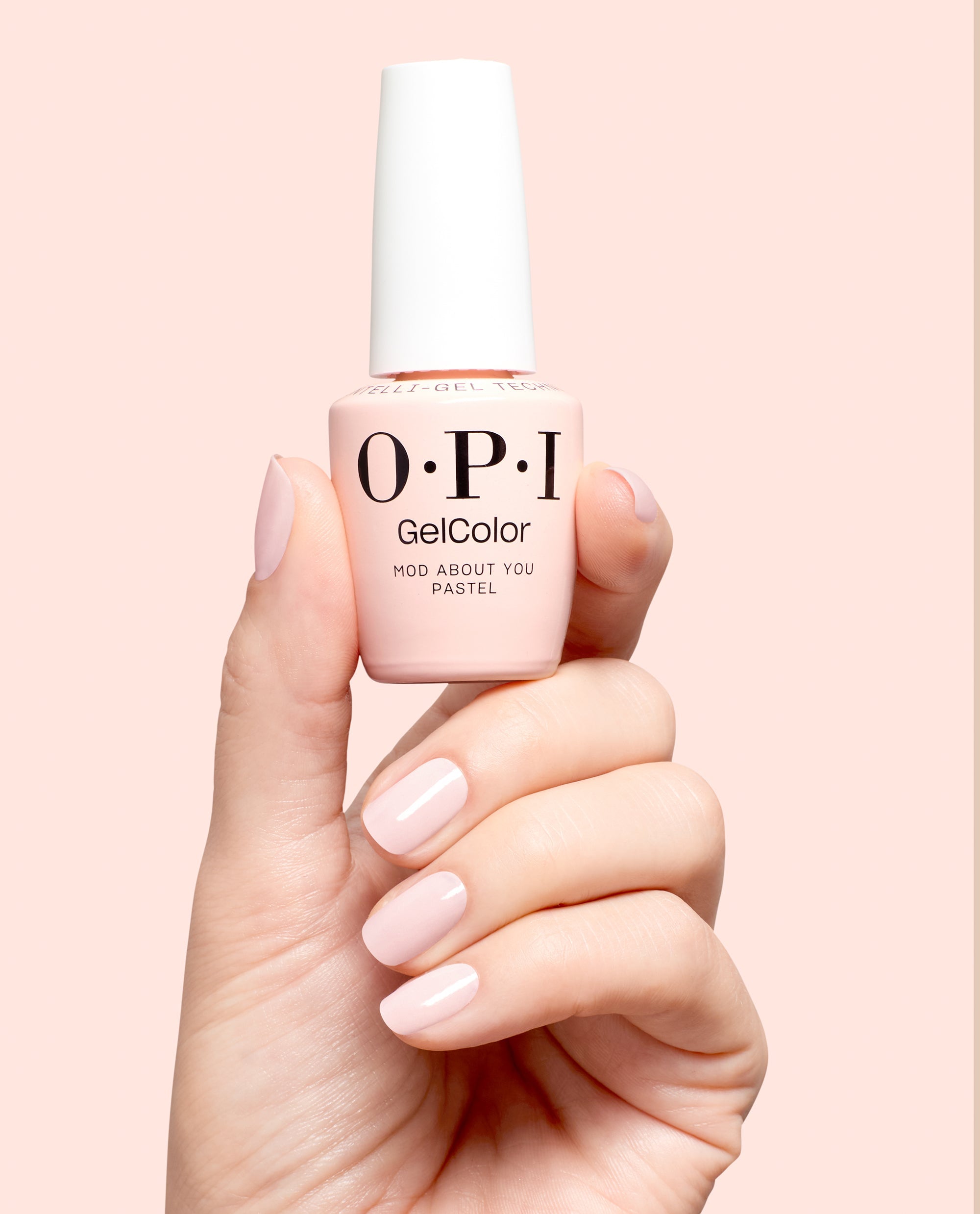 Hand holding OPI GelColor Mod About You Pastel gel nail polish with glossy pale pink manicured nails against a soft blush background, a delicate OPI shade for fall nail colors.