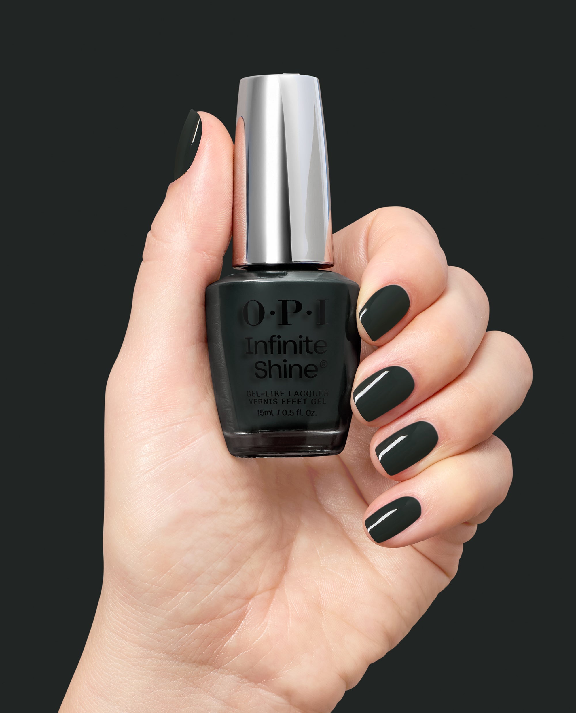 Hand holding OPI Infinite Shine Nein! Nein! Nein! OK Fine! nail lacquer, with glossy deep forest green nails showcasing a bold shade for fall nail colors against a dark background.