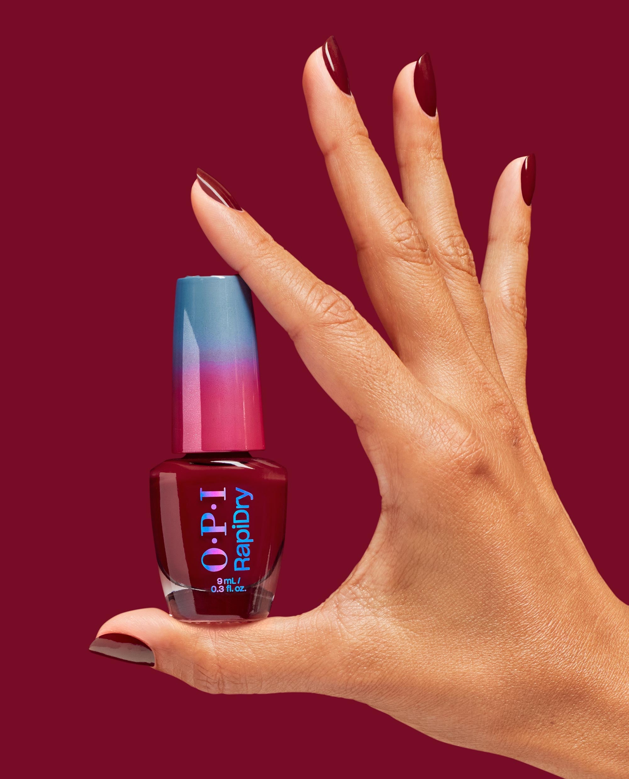 Hand with glossy deep beet-red nails holds OPI RapiDry Skip a Beet nail polish bottle with blue-pink cap against a burgundy background, showcasing rich fall nail colors.
