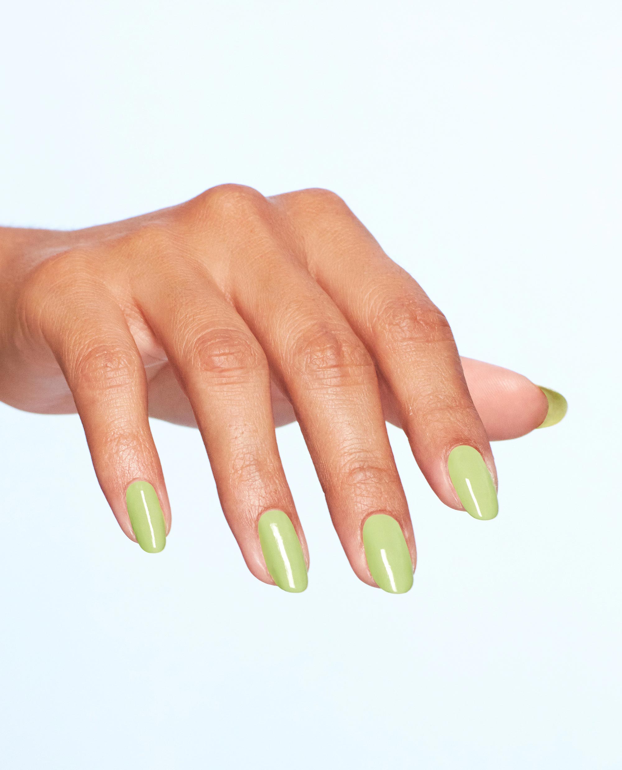 Manicured hand displays OPI Clear Your Cash nail lacquer, a glossy pastel lime green nail polish on medium-length oval nails, fanned outward against a clean light-blue studio background for fresh spring nail colors.