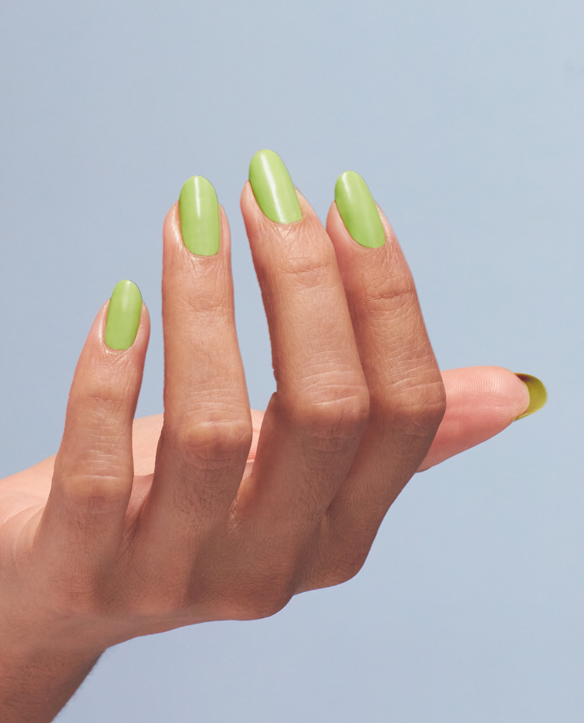 Manicured hand showcasing OPI Summer Monday-Fridays Nail Lacquer, a glossy light green nail polish, nails oval and neatly filed, fingers posed against a soft blue backdrop, spotlighting fresh spring nail colors for weekday wear.
