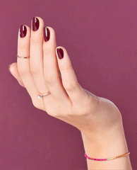 Hand with neatly manicured nails painted in deep burgundy Yes, My Condor Can-do! OPI nail polish, fingers slightly curved to show glossy coverage, against a matching plum background with delicate rings and a thin pink and gold bracelet.